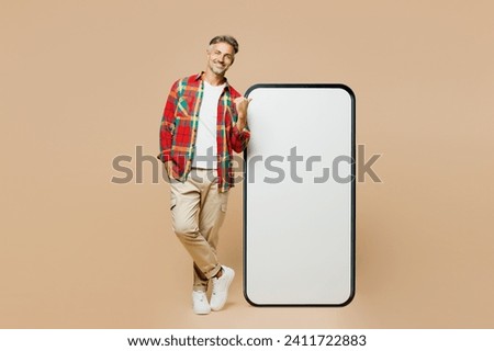 Full body adult man wear red shirt white t-shirt casual clothes point thumb finger on big huge blank screen mobile cell phone smartphone with area isolated on plain pastel light beige color background Royalty-Free Stock Photo #2411722883