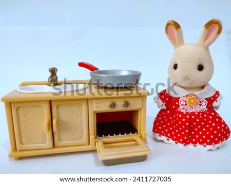An adorable rabbit characters toys wearing a red dress next to a mini sink, there is a frying pan on the stove and an open oven door with white background