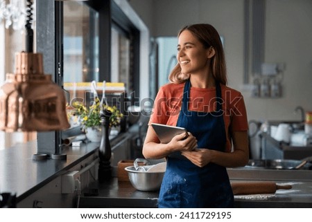 Young adult smiling female baker looking through window while standing in pastry kitchen and holding digital tablet. Royalty-Free Stock Photo #2411729195
