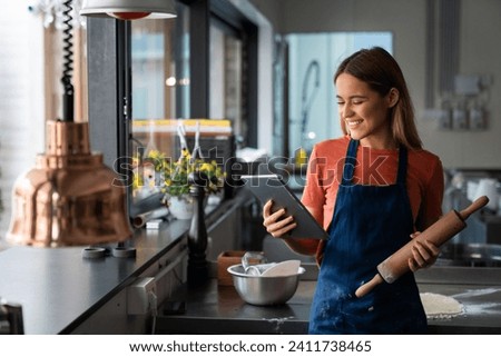 Cheerful female food blogger professional baker looking at digital tablet in commercial kitchen. Portrait of successful woman baker with digital tablet in pastry kitchen. Royalty-Free Stock Photo #2411738465