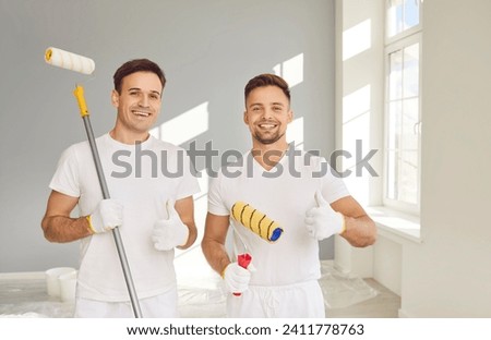 Portrait of smiling happy team of house painters looking at camera with paint rollers and showing thumb up sign. Building contractors paint the wall of new apartment doing repair renovation.