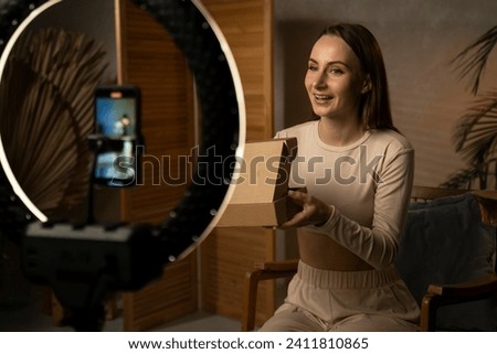 Young, beautiful, cheerful female blogger takes pictures on her phone camera as she unpacks a parcel from an online store and tells subscribers about buying goods while sitting at a table in her own