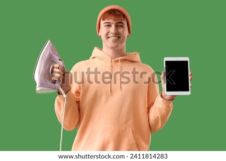 Handsome young man holding electric iron and tablet computer with blank screen on green background