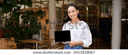 Smiling asian woman shows tablet screen and looks pleased, stands in cafe.