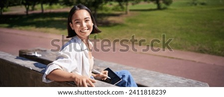 Happy korean woman sitting on bench with digital tablet and graphic pen, smiling, turning back at camera and talking lively.