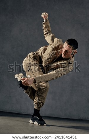 Attractive male with tattooed body, earrings, beard. Dressed in khaki jumpsuit and black sneakers. Dancing on gray background. Dancehall, hip-hop