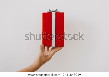 Picture of hand holding red gift box with silver ribbon, festival season, celebration, showing red present isolated on white background wall.