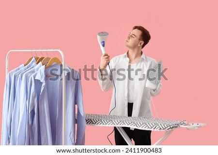 Doubtful young man with garment steamer and electric iron on pink background