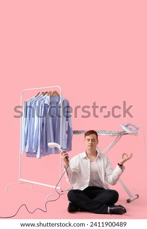 Handsome young man with garment steamer meditating near ironing board and clothes rack on pink background