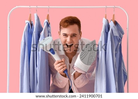 Excited young man with electric iron and garment steamer near clothes rack on pink background