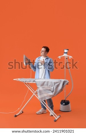 Handsome young man pointing at electric iron near ironing board on orange background