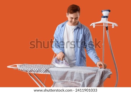 Handsome young man ironing clothes on orange background