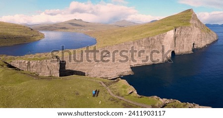 Aerial photography couple hiking and admiring the view Trælanípa, the famous slave cliff in Faroe Islands. Scenic scenery Leitsvatn, a lake in a different place with a seaside waterfall.
