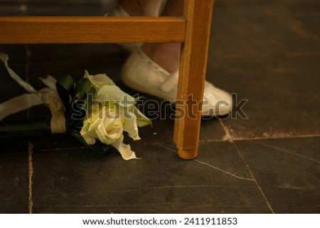 This photograph presents a poignant scene where a bridal bouquet of white roses lies forgotten on a dark wooden floor, juxtaposed against the background of a bride's white shoes. The image conveys a