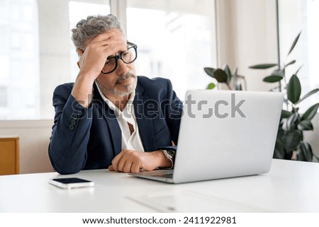 Thoughtful male office employee using laptop indoors, looking at laptop screen with puzzled face expression, businessman solving tasks, having problems with finance Royalty-Free Stock Photo #2411922981