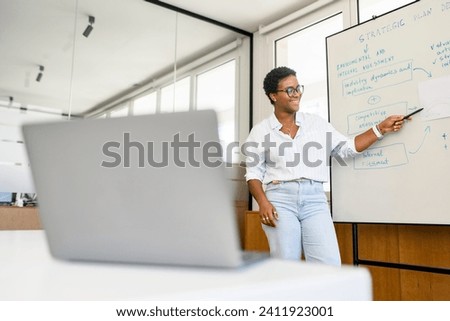 Young stylish African-American female business coach or speaker make flip chart presentation online, explaining project plan to online audience in front of the laptop during virtual conference
