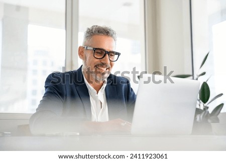 Smiling mature businessman wearing formal suit and glasses using laptop for office work, male employee looking at the screen, reading news, checking mail Royalty-Free Stock Photo #2411923061
