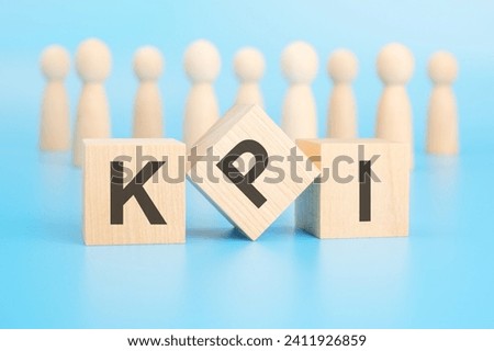 forming a conceptual word with blocks in the foreground - KPI (Key Performance Indicators). the blocks are located on a blue surface in front of wooden figures symbolizing people. Royalty-Free Stock Photo #2411926859