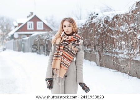 Beautiful cute blonde girl in a fashion beige coat with a vintage style scarf and mittens walks in the countryside on a snowy winter day