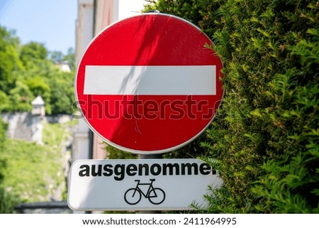 A red round sign with a white bar in the middle, which means that you are not allowed to drive into the road. Below the sign is a sign that bicycles are excluded. The signs are next to a green bush