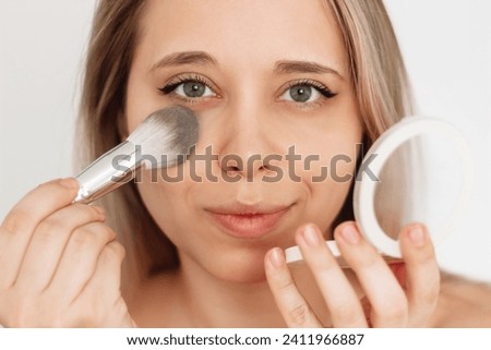Close up of a young caucasian blonde woman using a brush for applying of colorless face powder to the skin under her eyes on a white background. A girl doing makeup. Cosmetic beauty concept Royalty-Free Stock Photo #2411966887