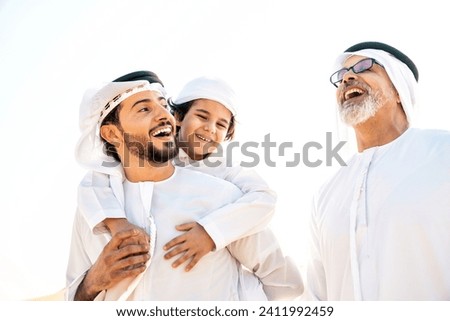 Three generation family making a safari in the desert of Dubai wearing white kandura outfit. Grandfather, son and grandson spending time together in the nature. Royalty-Free Stock Photo #2411992459