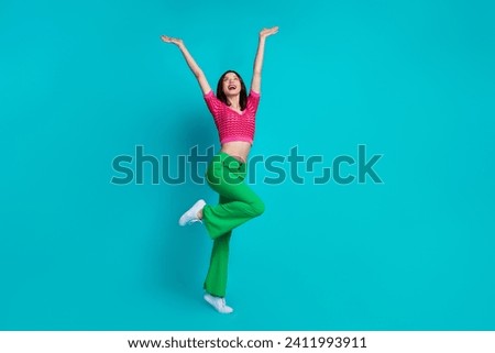 Full size photo of overjoyed girl wear pink top retro trousers raising palms up look empty space isolated on blue color background