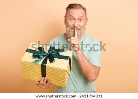 Photo of positive aged man hand hold giftbox finger touch lips demonstrate shh do not talk gesture isolated on beige color background