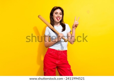 Photo of funny carefree woman with brunette hair dressed t-shirt showing tongue v-sign hold bat isolated on yellow color background