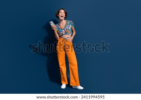 Full size photo of attractive young woman curious empty space gadget dressed stylish flared pants isolated on dark blue color background