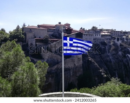 Flag of Greece is displayed against background of ancient monastery located on impregnable rock. Blue sky on bright sunny day. Selective focus. Copy space.