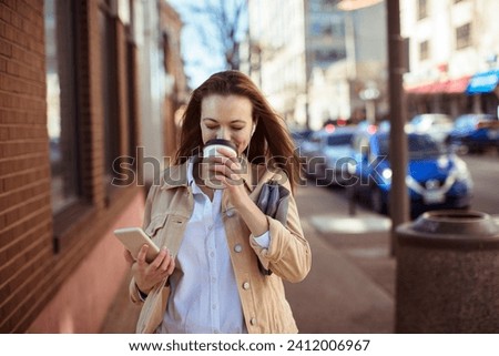 Smiling woman walking with coffee and looking at smartphone on city street