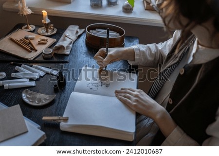 Woman paintress improving painting skills creating sketches in sketchbook sitting at desk with candle. Art worker girl artist freelancer illustrator practicing drawing enjoying process in workshop. 