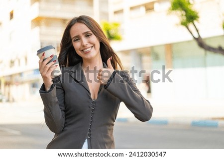 Young woman holding a take away coffee at outdoors with thumbs up because something good has happened