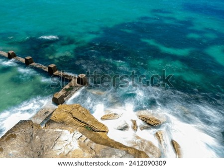 Powerful white swirling waves (blurred motion) crashing into rocky coastline. Public recreational rockpool to left. Ocean water showing tropical blue and green colors of the shallow floor.