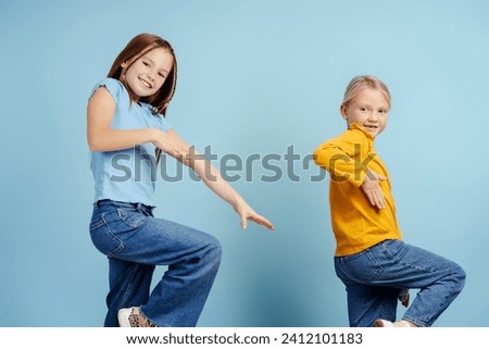 Happy girls, cute sisters looking at camera and dancing isolated on blue background. Young happy fashion models posing for pictures in studio. Childhood, positive lifestyle concept 