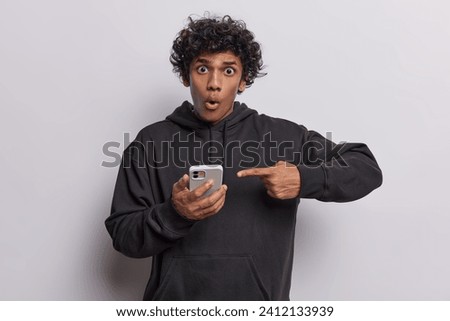 Shocked curly haired Hindu man points index finger on smartphone gets amazing news receives message wears casual black sweatshirt isolated over white background. Didital communication concept