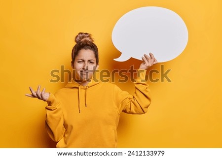 Hesitant puzzled young woman with hair bun shrugs shoulders holds blank communication bubble for your advertising content doesnt know what to write here isolated over vivid yellow background Royalty-Free Stock Photo #2412133979
