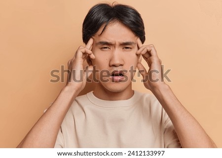 Photo of displeased Chinese man keeps hands on temples suffers from headache massages temples tries to concentrate or remember something dressed in casual t shirt isolated over brown background.