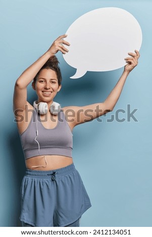 Vertical shot of positive young European sporty woman dressed in activewear poses with headphones around neck holds blank speech bubble for your promotional text isolated over blue background