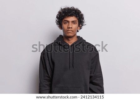Waist up shot of serious curly haired Hindu man looks directly at camera has confident gaze calm expression dressed in casual black sweatshirt isolated over white background has important conversation