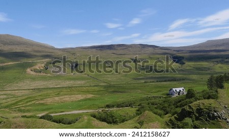 A lone house stands embraced by the vastness of green, rolling hills under a blue sky, dotted with white clouds. Shadows play across the terrain, hinting at the sun's position. Royalty-Free Stock Photo #2412158627