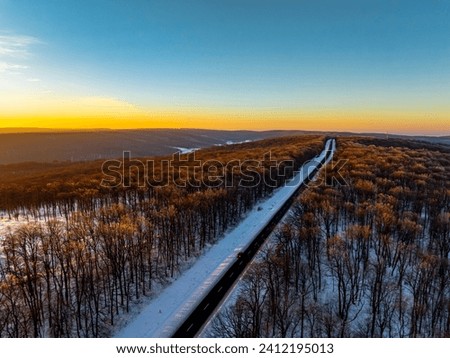 aerial view over the clean road in the forest with the treetops illuminated by the setting sun during winter sunset