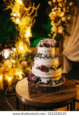 A three-tiered wedding cake adorned with fresh flowers and greenery, set against a backdrop of warm candlelight. Royalty-Free Stock Photo #2412230135