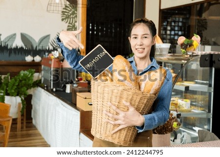 Asian young barista worker female holding Promotion sign board and bread in basket in front of bakery cabinet taking pictures to promote in social media channel, happy small business owner lifestyle