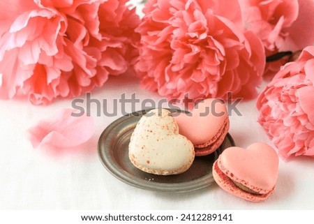 Pink coral peony flowers and french sweet cookies macarons macaroons on linen table cloth background.  Heart shape macarons and bouquet of peony. Pastry shop love or birthday card with copy space.