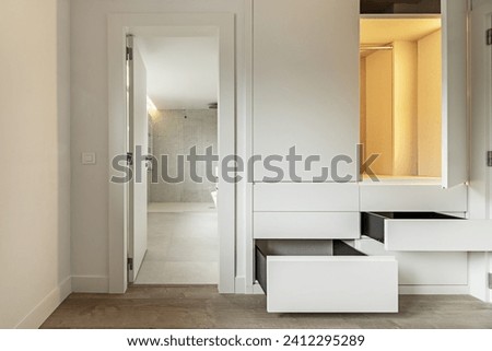A bedroom with access door to a private bathroom and built-in wardrobe with drawers and open wooden doors with interior LED lights on Royalty-Free Stock Photo #2412295289