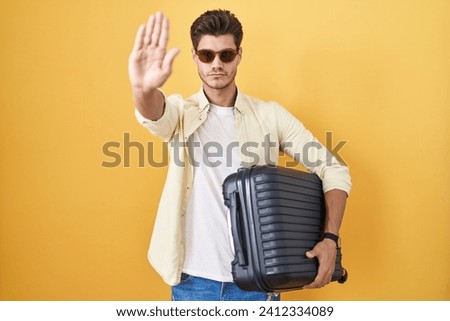 Young hispanic man holding suitcase going on summer vacation doing stop sing with palm of the hand. warning expression with negative and serious gesture on the face. 
