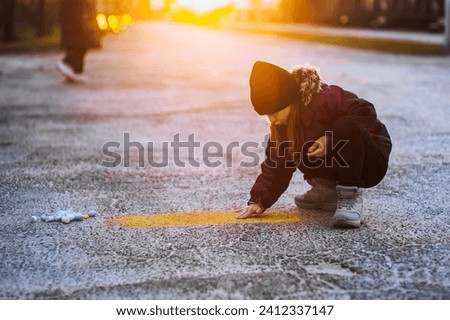 A little diligent lonely girl, a child draws a drawing with colored chalk on the asphalt on the street in the park at sunset. Photography, close-up portrait, design, childhood concept.