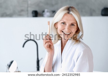 Cheerful mature woman in white bathrobe hold wooden toothbrush in hand and smile at home bathroom. Female using zero waste brush for cleansing teeth. Oral hygiene concept Royalty-Free Stock Photo #2412352985